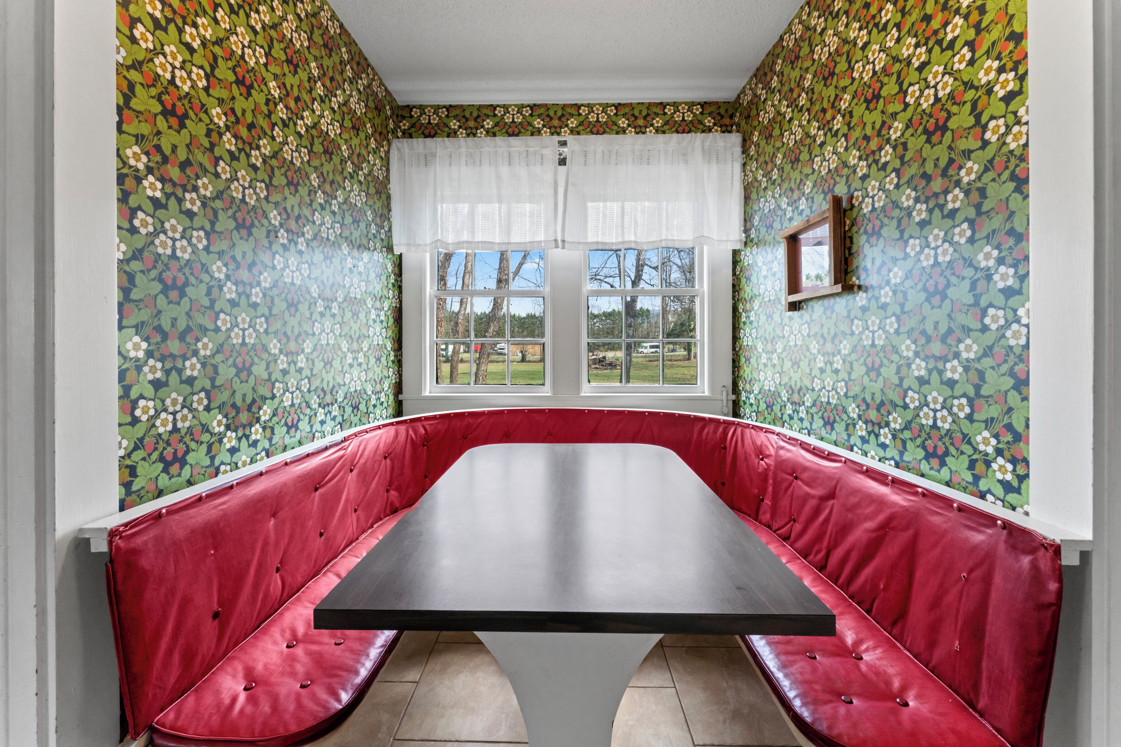 A room with a red cushioned bench, a large table, and a window with a view of trees and a lawn.