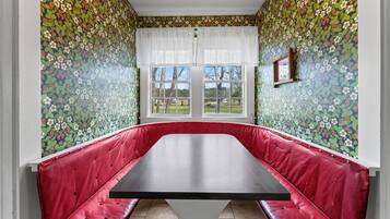 A room with a red cushioned bench, a large table, and a window with a view of trees and a lawn.