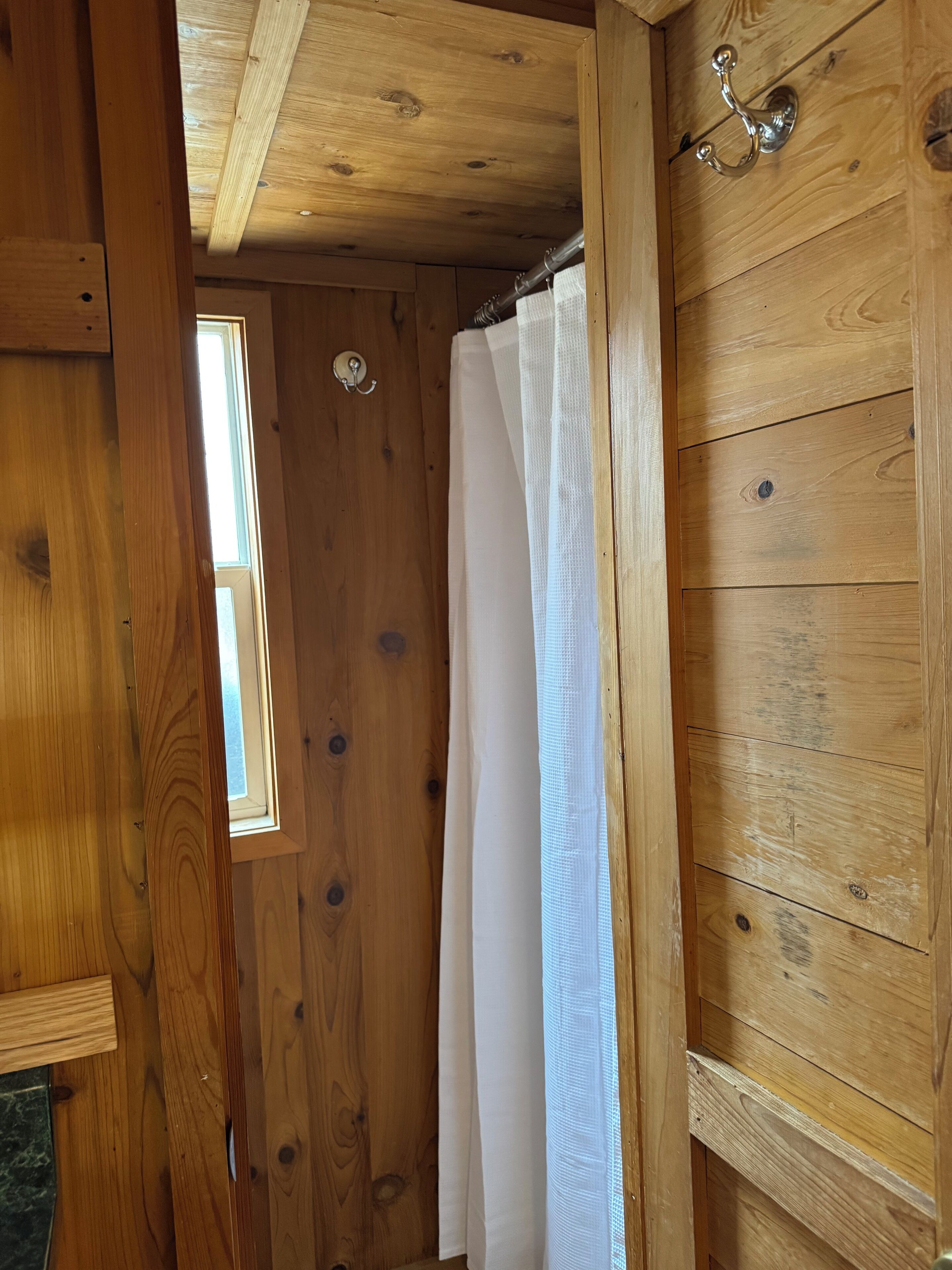 A wooden interior with a window, a white curtain, and a metal hook on the wall.