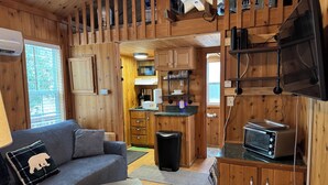A cozy cabin interior with a living area featuring a sofa and a small table, a kitchen with wooden cabinets and appliances, and a television mounted on the wall. - Tiny Home II, Pet Friendly, Free Cancellations (Boone)