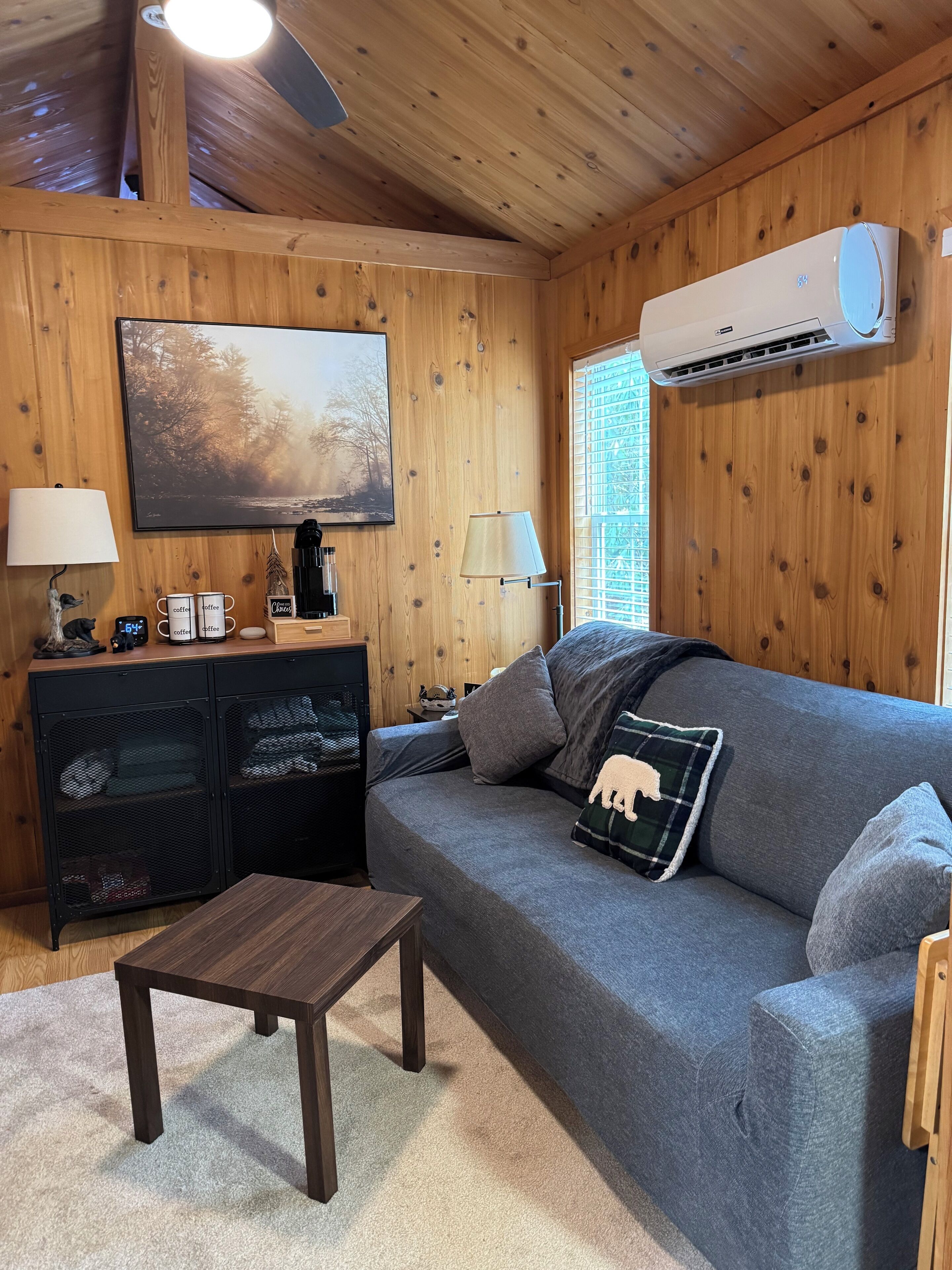 A cozy living room with a grey sofa, a small wooden table, a coffee station, and a painting on the wall.