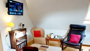 Uma sala de estar aconchegante com piso de madeira, uma televisão montada na parede, uma estante de livros, uma pequena mesa com um tabuleiro de xadrez e duas cadeiras com almofadas vermelhas.