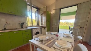 A compact kitchen with green cabinets, a built-in washing machine, and a dining area with a table set for four. - Apartment in a Villa With a Relaxing Pool (Ispica)