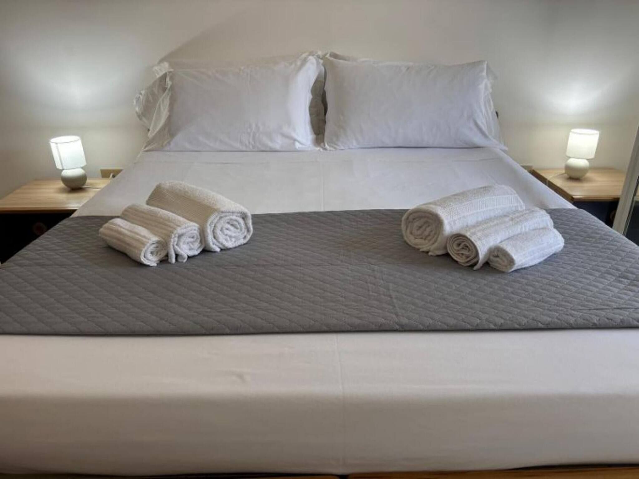 Una cama bien hecha con sábanas blancas y dos toallas enrolladas sobre la cama. Hay dos mesitas de noche con lámparas a cada lado de la cama.