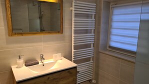 Ein modernes Badezimmer mit einem hölzernen Waschtisch, einer Duschkabine aus Glas, einem weißen Handtuchheizkörper und einem Fenster mit Jalousien.