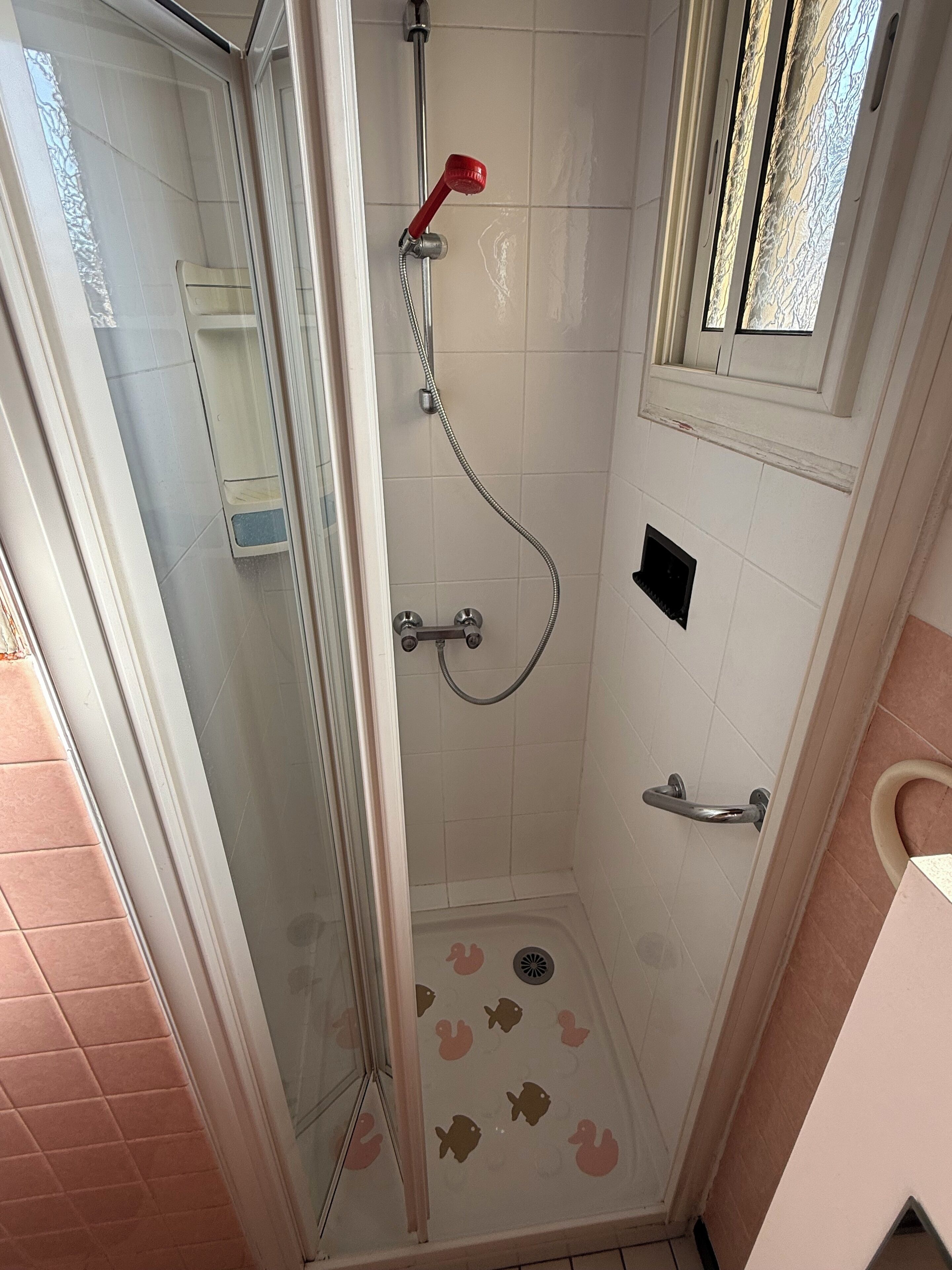 A shower with a glass door, a red shower head, and a handrail.
