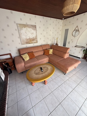 A living room with a sectional sofa, a round coffee table, and a large painting on the wall. - Cosy house with garden - near Mimizan beach (Mimizan)
