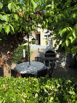 A patio with a round table and chairs, a stone barbecue, and a building in the background. - Cosy house with garden - near Mimizan beach (Mimizan)