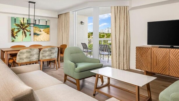 Un salon moderne comprenant un canapé, des fauteuils, une table à manger avec des chaises, un téléviseur et un balcon offrant une vue sur les immebles et les palmiers.