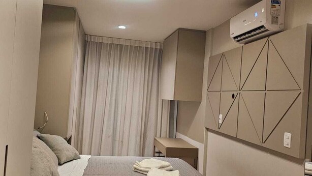A modern hotel room with a neatly made bed, a bedside table, a lamp, and an air conditioning unit mounted on the wall. - Apto 2 Quartos com 1 Suíte a Poucos Passos da Praia (Vila Velha)