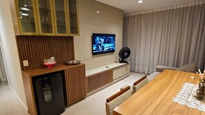 A modern kitchen with a built-in dishwasher, a dining area with a wooden table and chairs, and a TV mounted on the wall. - Apto 2 Quartos com 1 Suíte a Poucos Passos da Praia (Vila Velha)