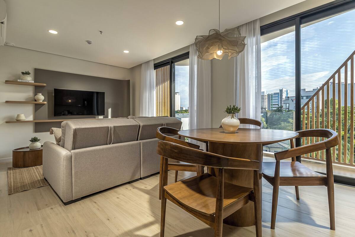 A modern living room with a sofa, TV, dining table, chairs, and large windows.
