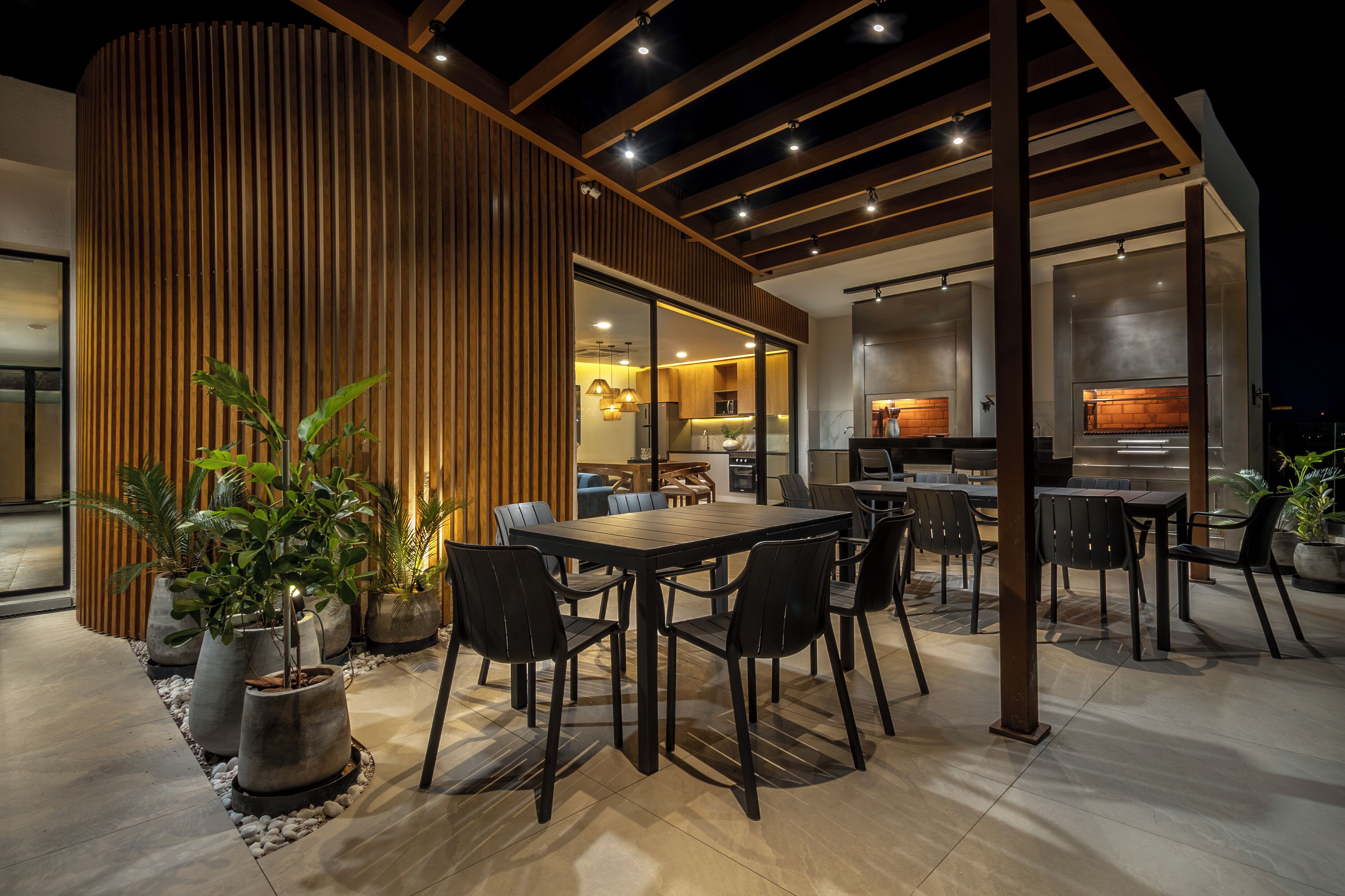 An outdoor dining area with a wooden pergola, potted plants, and a modern kitchen visible through the glass doors.