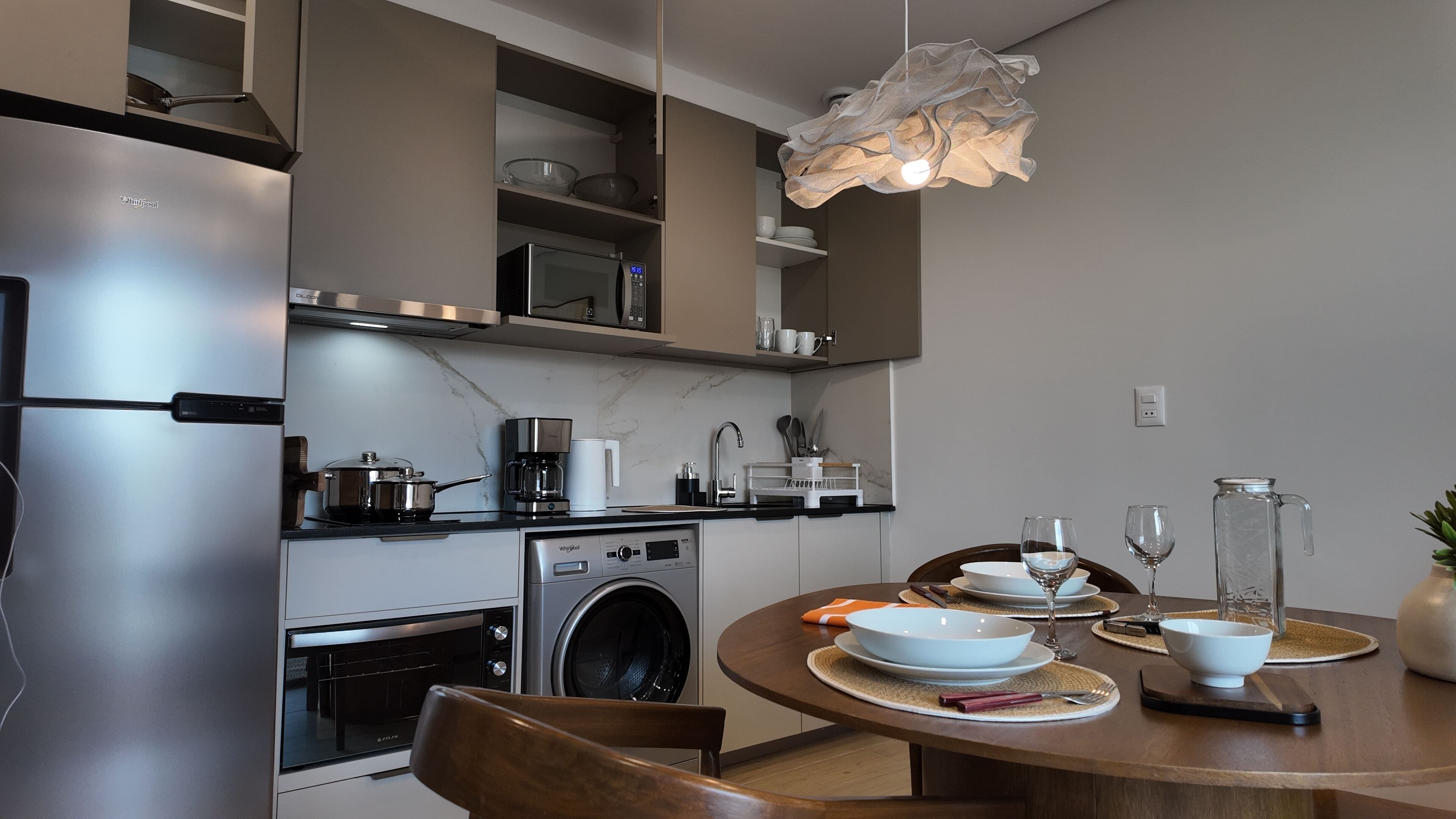 A modern kitchen with a stainless steel refrigerator, a built-in oven, a microwave, a washing machine, and a dining area with a round table set for two.