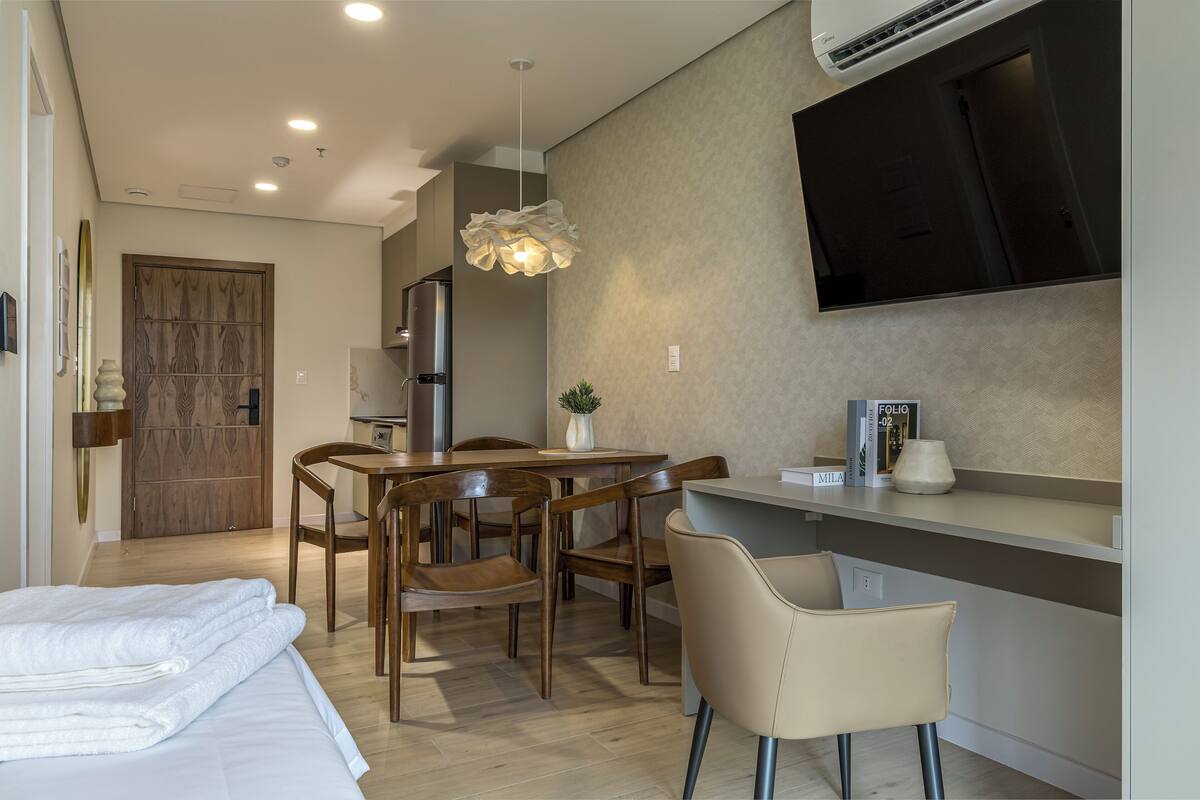 A modern hotel room with a bed, a dining area with chairs and a table, a TV mounted on the wall, and an air conditioning unit.