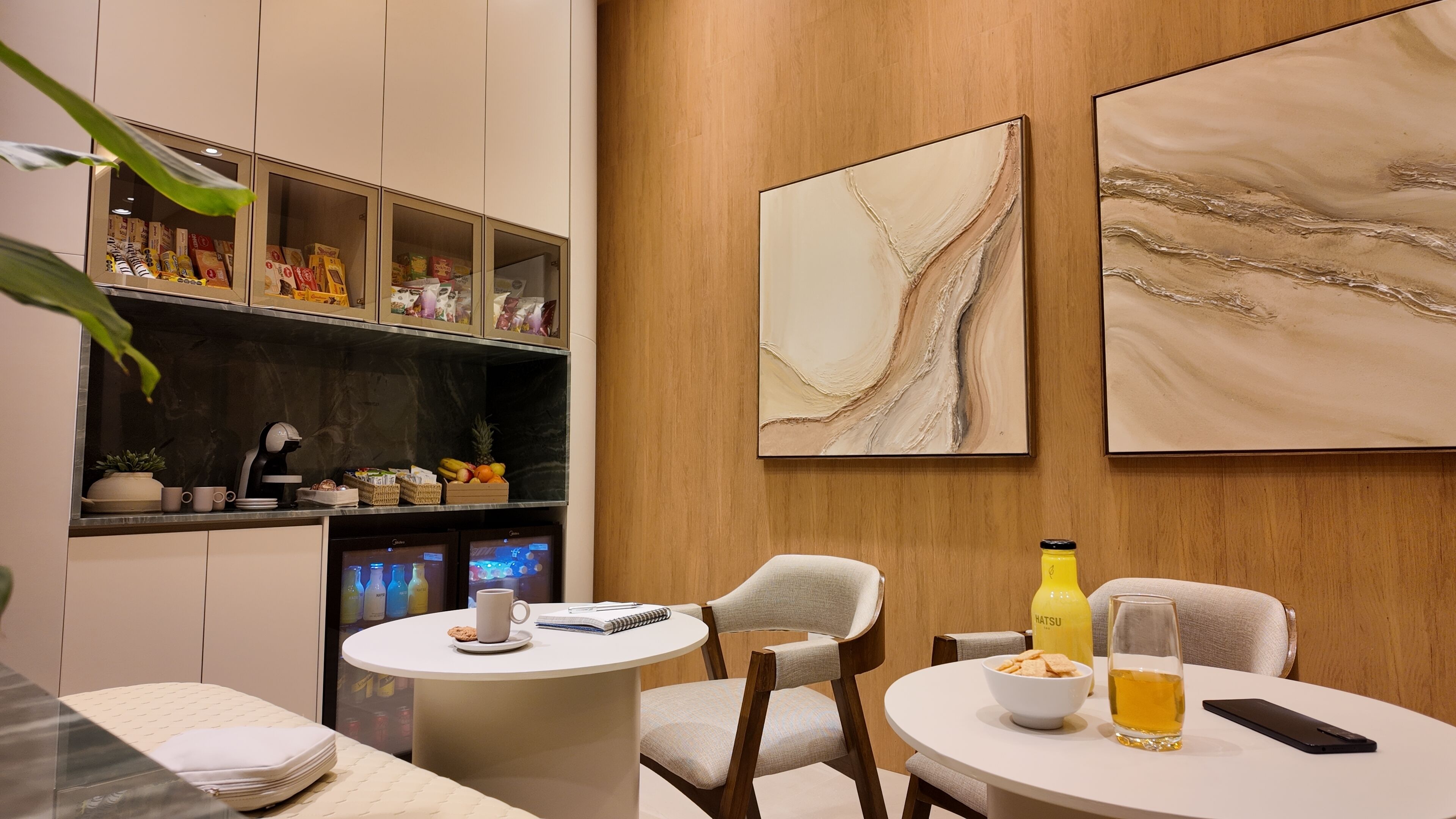 A modern hotel room with a minibar, two round tables with chairs, a coffee machine, and artwork on the walls.