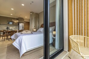 A hotel room with a bed, a desk with a chair, a kitchenette, and a view through a glass door.