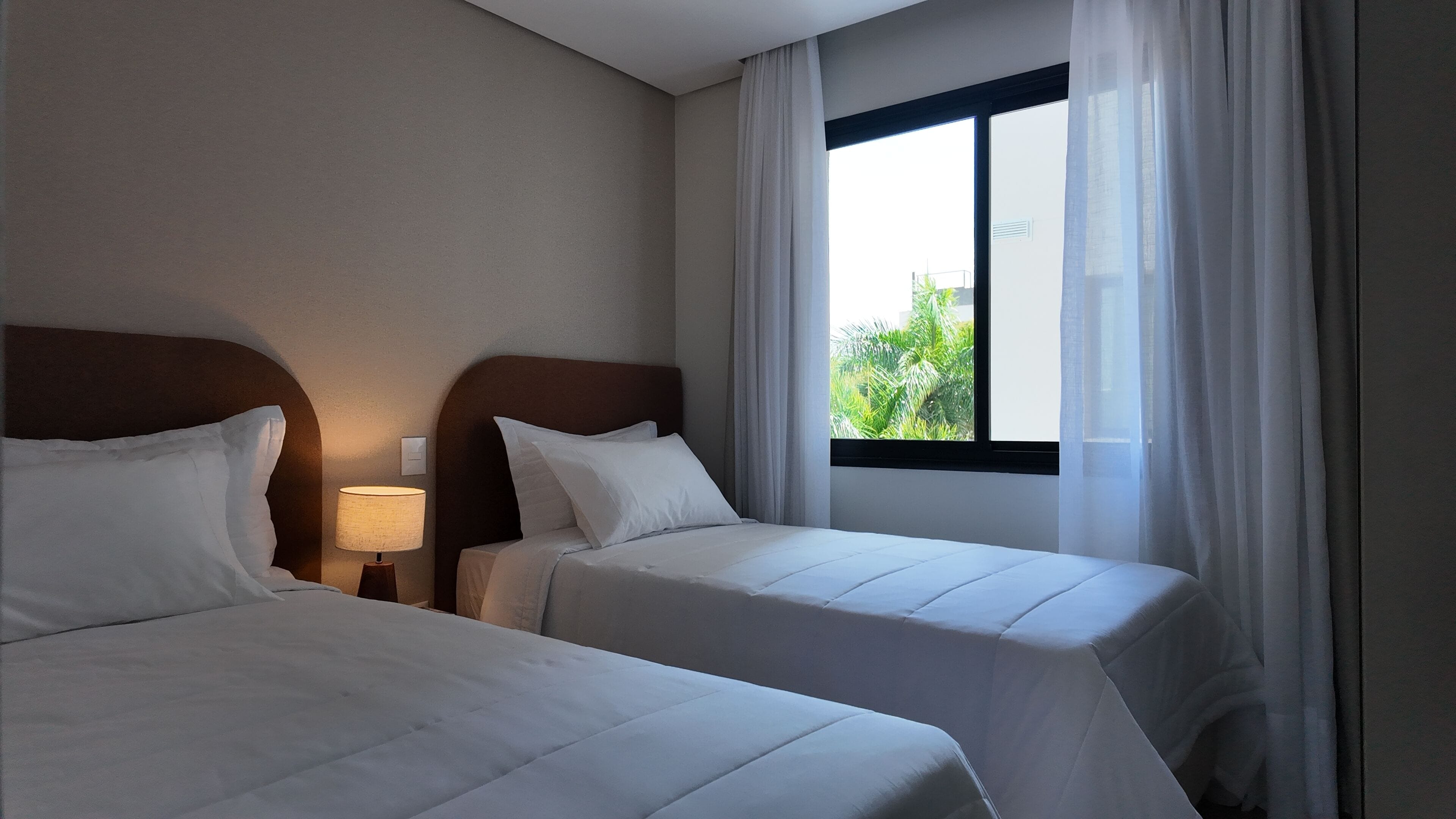 Une chambre d’hôtel avec deux lits, une table de chevet avec une lampe, une fenêtre avec des rideaux et une vue sur la verdure.