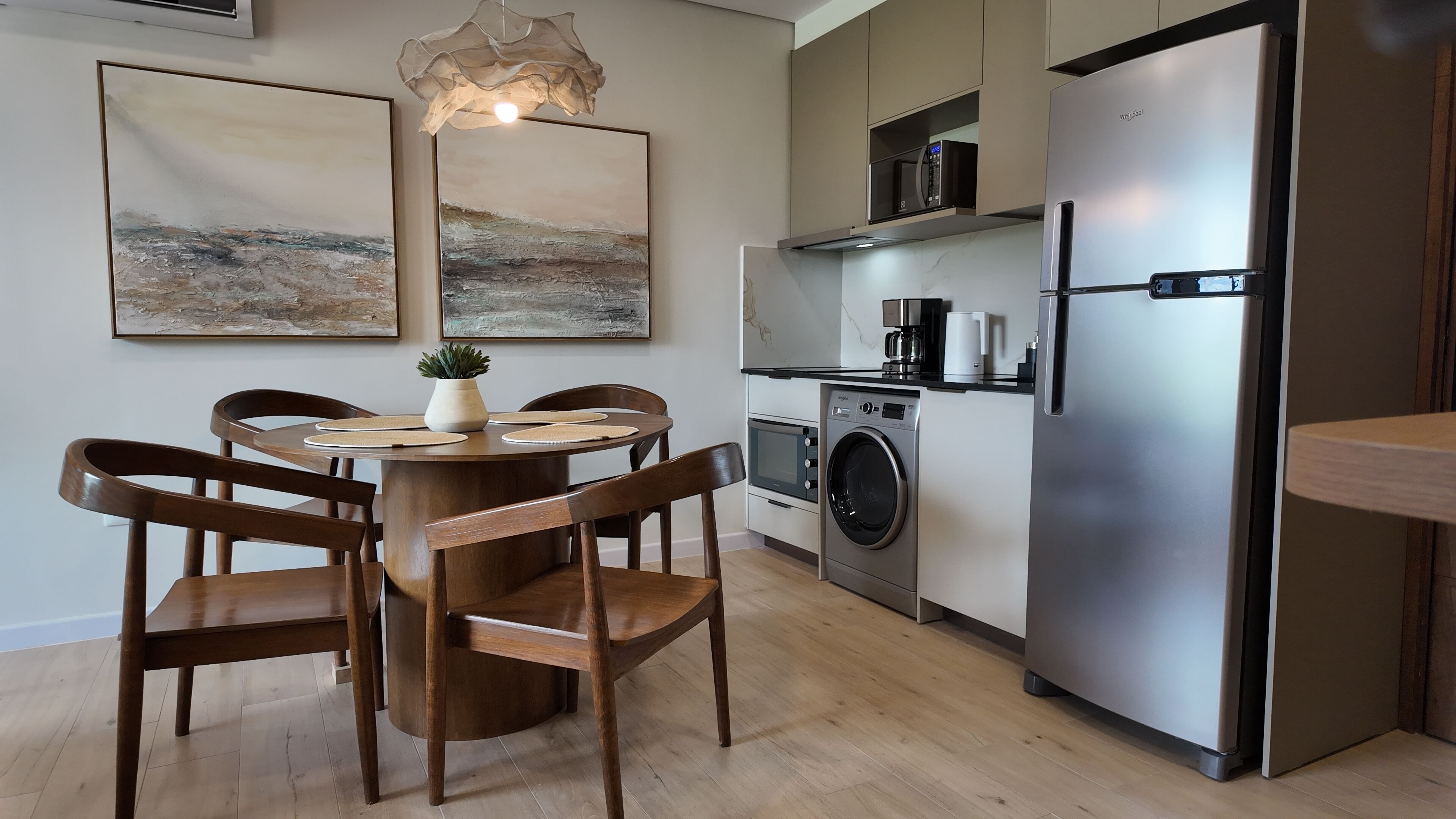 A modern kitchen with a stainless steel refrigerator, a built-in oven, and a washing machine. There is a dining table with chairs and two framed artworks on the wall.