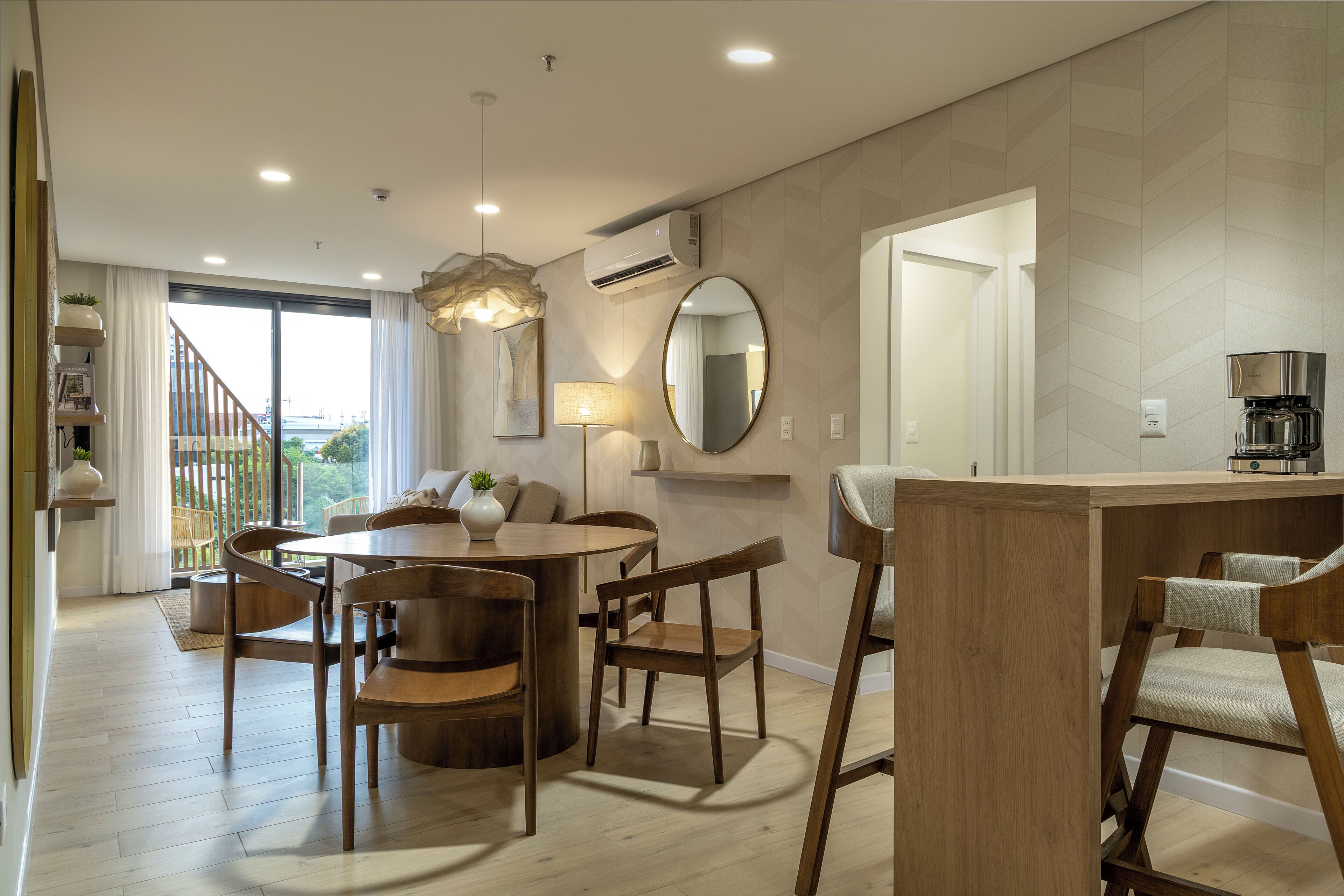 A modern dining area with a round table, wooden chairs, a hanging light fixture, and a view through a glass door.