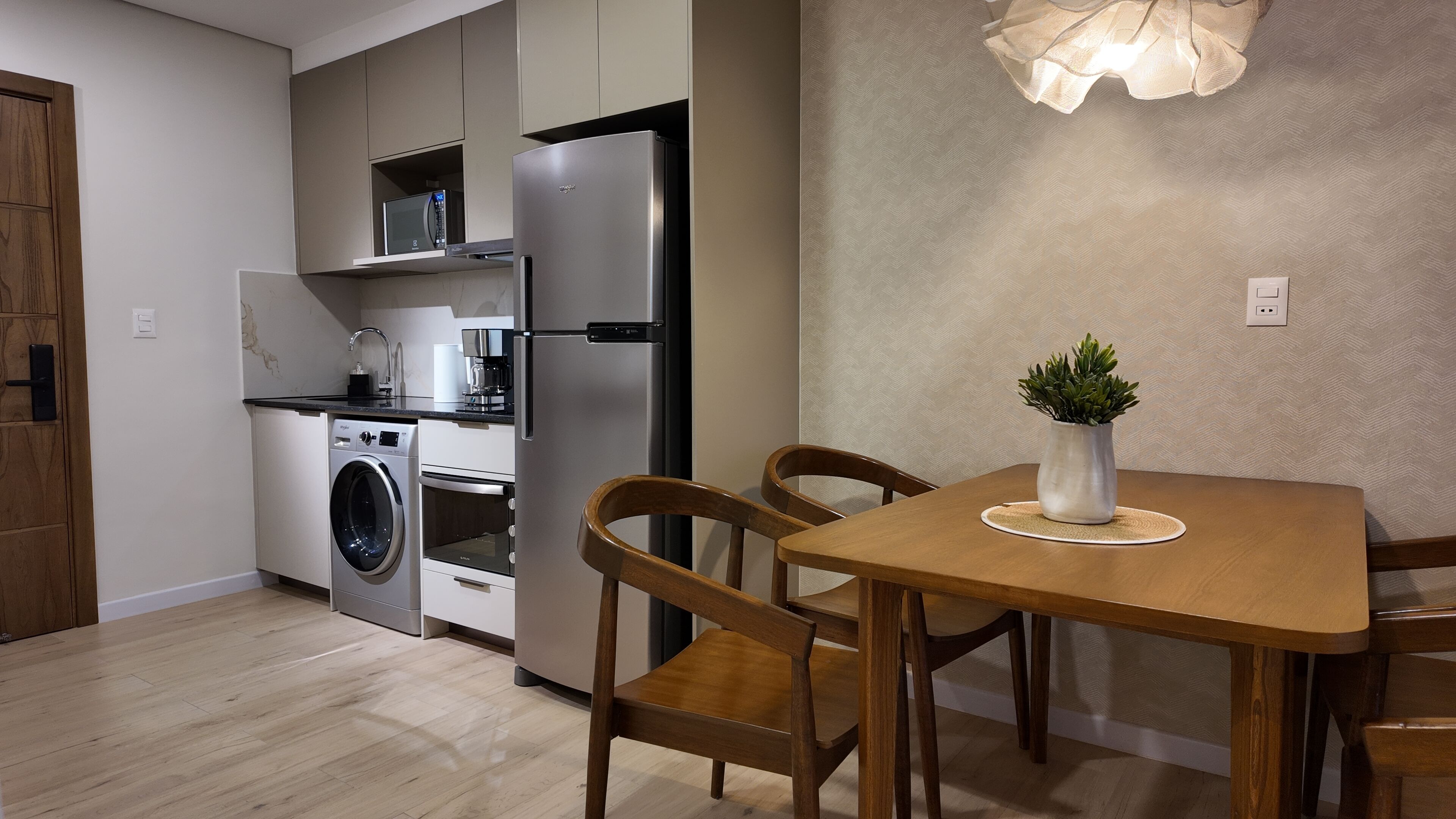 A compact kitchen with a washing machine, oven, and refrigerator. There is a dining table with chairs and a potted plant on it.
