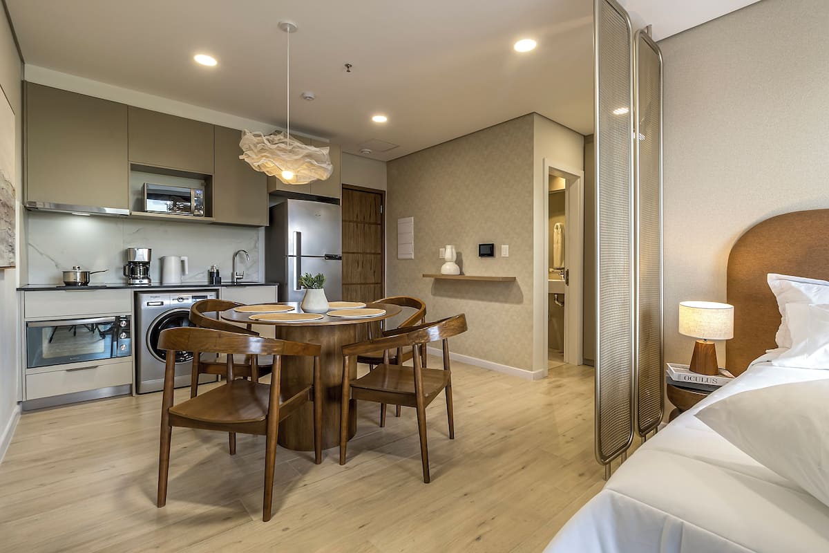 A modern hotel room with a bed, a bedside table with a lamp, a dining area with chairs and a table, a kitchen with appliances, and a shower area.