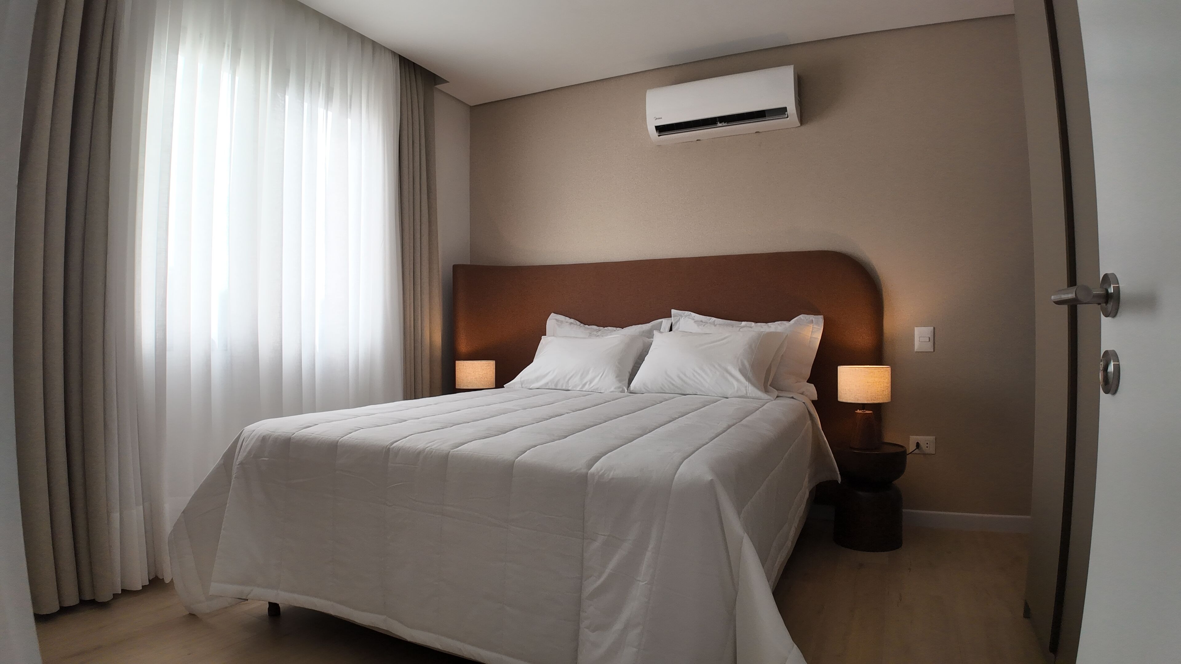 Hotellihuone, jossa on suuri sänky, kaksi yöpöytävalaisinta, ilmastointilaite ja näkymä ulos ohuiden verhojen läpi.
