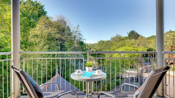 Un balcon avec une table et des chaises, une balustrade, et une vue sur la verdure et une route.