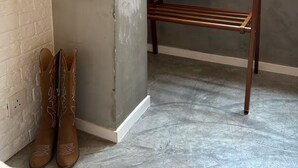 Ein Zimmer mit Betonboden, einer hölzernen Bank und einem Paar brauner Stiefel vor einer Ziegelwand.