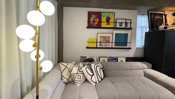 Ein modernes Wohnzimmer mit einer großen, L-förmigen Couch, einer Stehlampe mit mehreren Leuchten und einer weißen Ziegelwand mit gerahmten Kunstwerken.