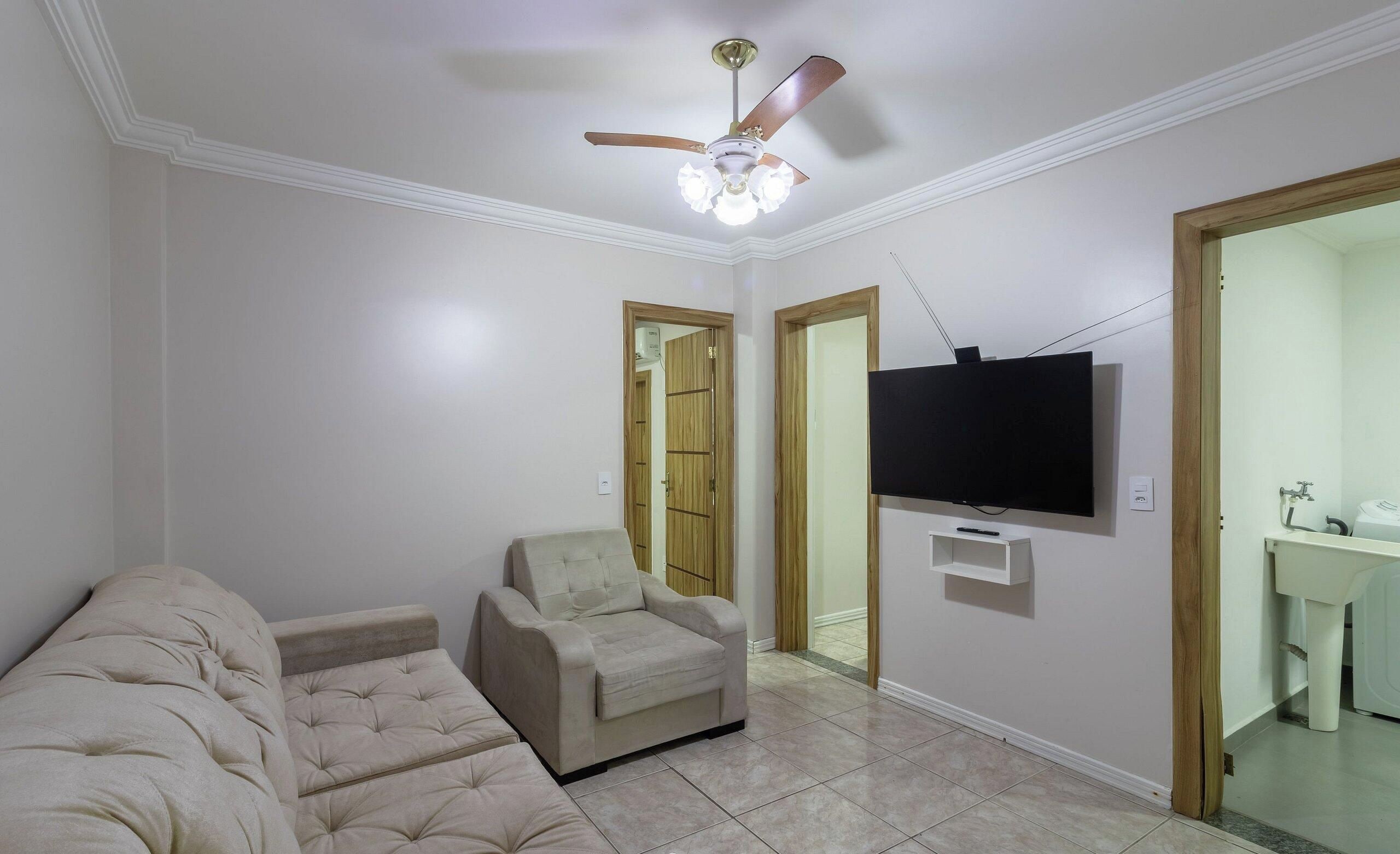 A living room with a sofa, a chair, a ceiling fan, and a television mounted on the wall.