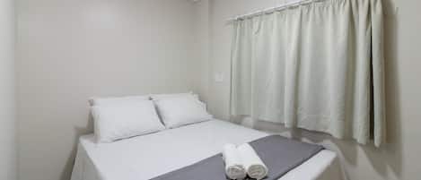 A neatly made bed with white linens and a grey blanket, a window with white curtains, and a wall-mounted light fixture.