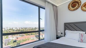 A bedroom with a large window, a bed with white bedding, a grey headboard, and two wall-mounted decorative plates. - Luxurious apartment near mall and Carrefour, giving a cozy and homely vibe (Nairobi)