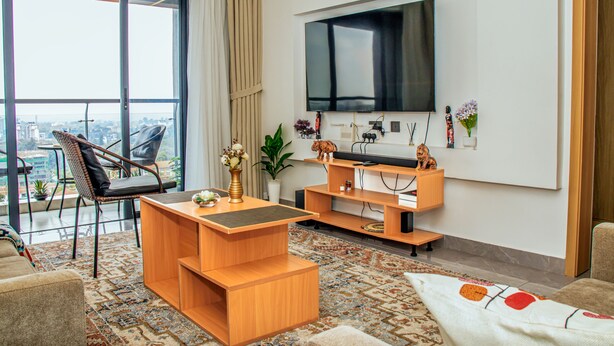 A modern living room with a large TV, wooden furniture, a view of the city, and a balcony. - Luxurious apartment near mall and Carrefour, giving a cozy and homely vibe (Nairobi)