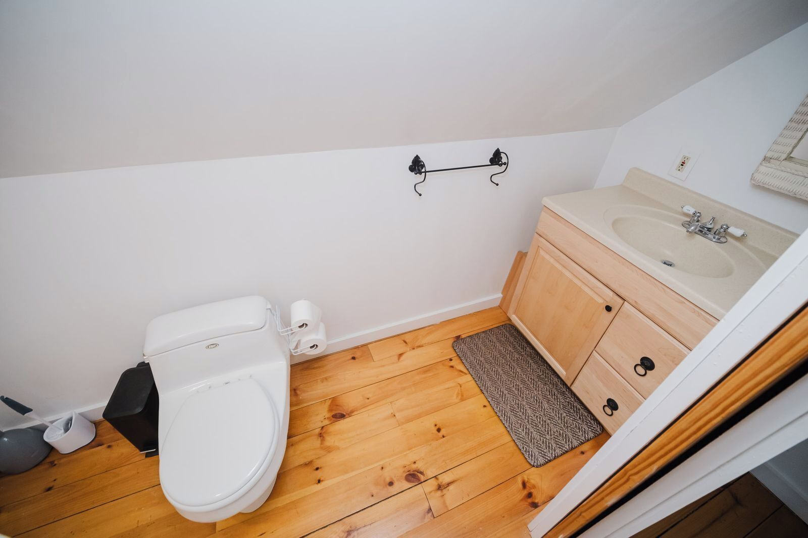 Une petite salle de bain propre, avec des toilettes blanches, un plancher en bois, un meuble en bois avec un lavabo et un porte-serviettes fixé au mur.