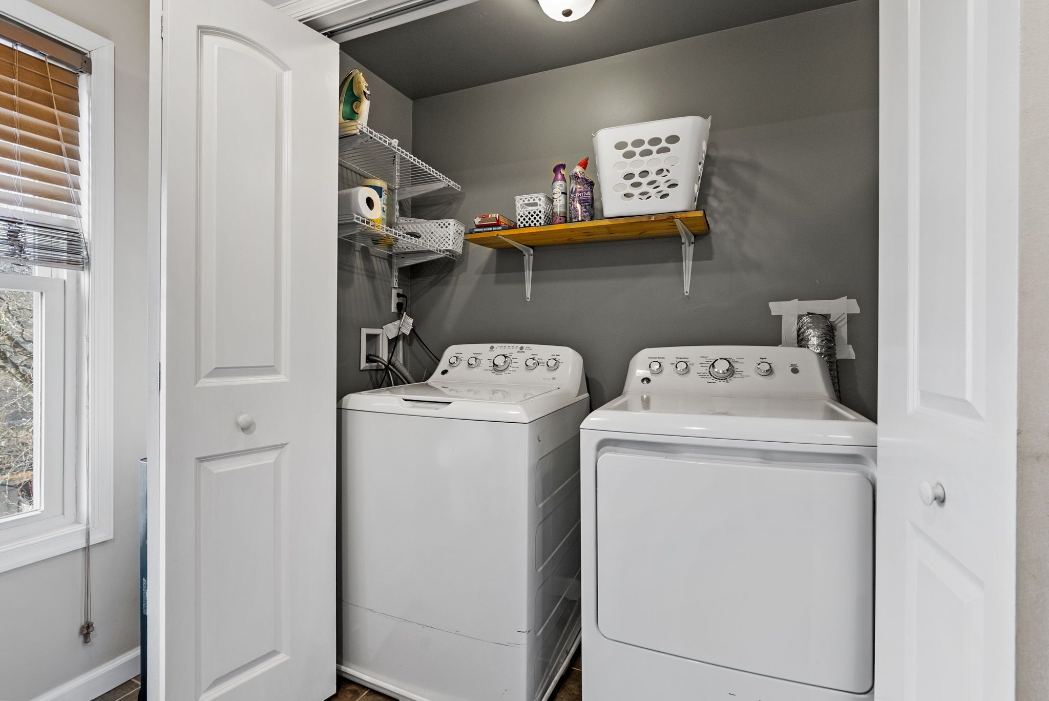 A laundry room with two white front-loading washers and dryers, a shelf with laundry supplies, and a window with blinds.