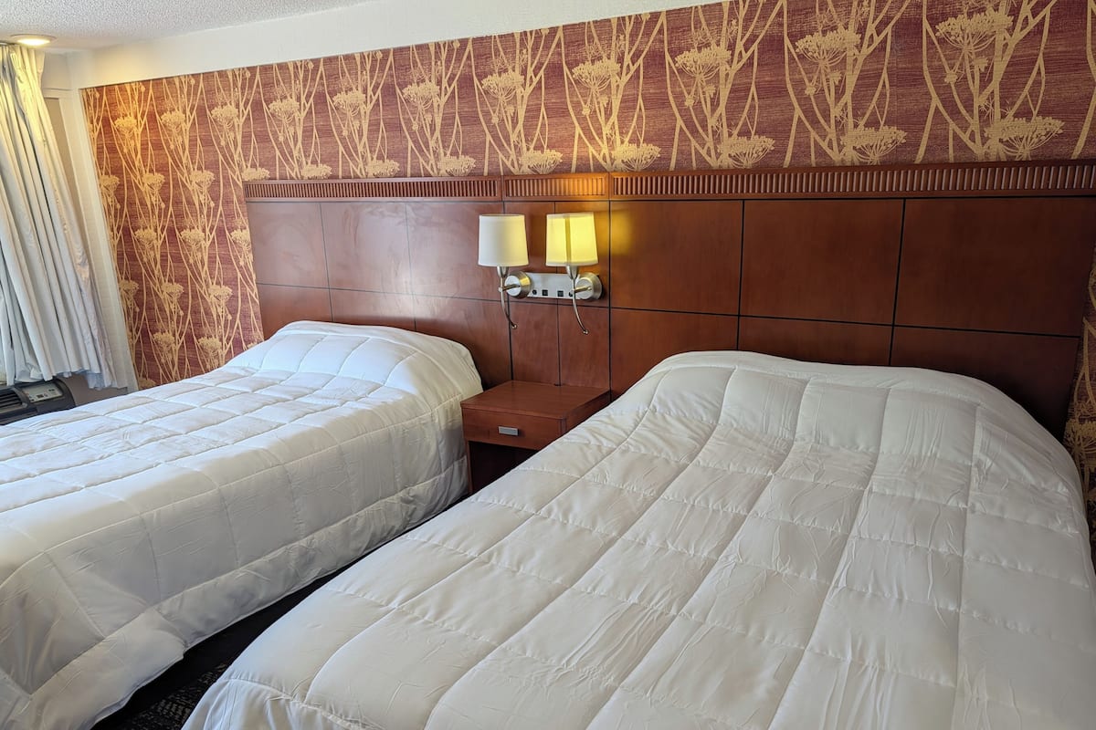 A hotel room with two beds, a wooden headboard, a nightstand with lamps, and a wall with a floral pattern.