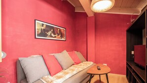 Una habitación con una pared roja, un sofá con cojines grises y rojos, una mesita de madera y un cuadro enmarcado sobre el sofá.