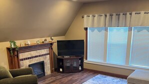 A living room with a fireplace, a flat-screen TV, a green armchair, a beige sofa, and a large window with blinds. - The Fannie Historic Apartment (Eau Claire)