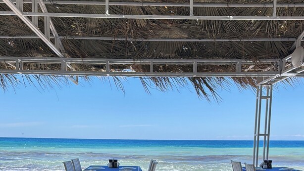 Un restaurant situé en bord de mer, avec des tables et des chaises disposées sous un toit de chaume, offrant une vue sur l’océan.