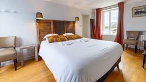 A hotel room with a large bed, wooden headboard, two bedside lamps, a small table, a chair, and a view of the outdoors through a window. - Cyrano & Roxane - 1 km de la plage (Ver-sur-Mer)