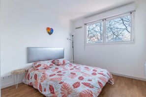 A bedroom with a bed, a bedside table, a lamp, and a window with a view of trees. - Le Petit Logis du Stade de France (Saint Denis)