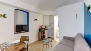 Ein modernes Wohnzimmer mit einem Holzesstisch, einer Couch, einem an der Wand montierten Fernseher und einer Küche im Hintergrund.