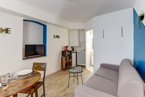 Un salon moderne avec une table à manger en bois, un canapé, un téléviseur fixé au mur et une cuisine en arrière-plan.