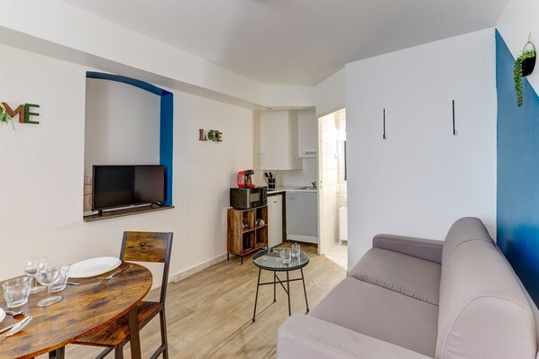 Ein modernes Wohnzimmer mit einem Holzesstisch, einer Couch, einem an der Wand montierten Fernseher und einer Küche im Hintergrund.