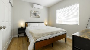 A bedroom with a wooden bed, white bedding, a nightstand with a lamp, a wall-mounted air conditioner, and a window with blinds.