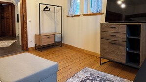 Une pièce avec un plancher en bois, un canapé gris, un meuble en bois doté de tiroirs, un téléviseur posé sur un meuble, et une fenêtre ornée de rideaux à motifs.