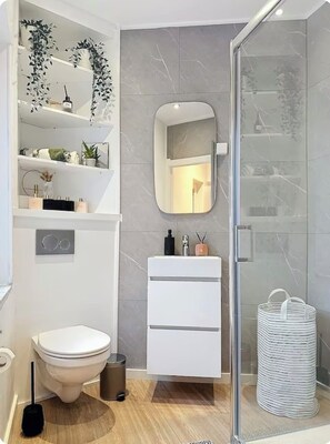 Une salle de bain moderne équipée de toilettes, d’un lavabo et d’une zone de douche. On y trouve des étagères avec des articles de toilette et un miroir au-dessus du lavabo.