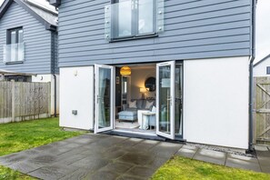 Uma casa moderna com fachada cinzenta, parte inferior branca e porta de vidro que dá para uma sala de estar com sofá e mesa.
