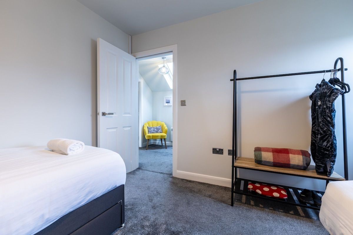 A bedroom with a bed, a bench with hanging clothes, and a door leading to another room.